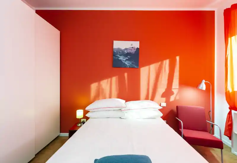 Dormitorio moderno y luminoso con cama doble centrada frente a una pared roja intensa, sillón acogedor, lámpara de noche y luz natural de la ventana.