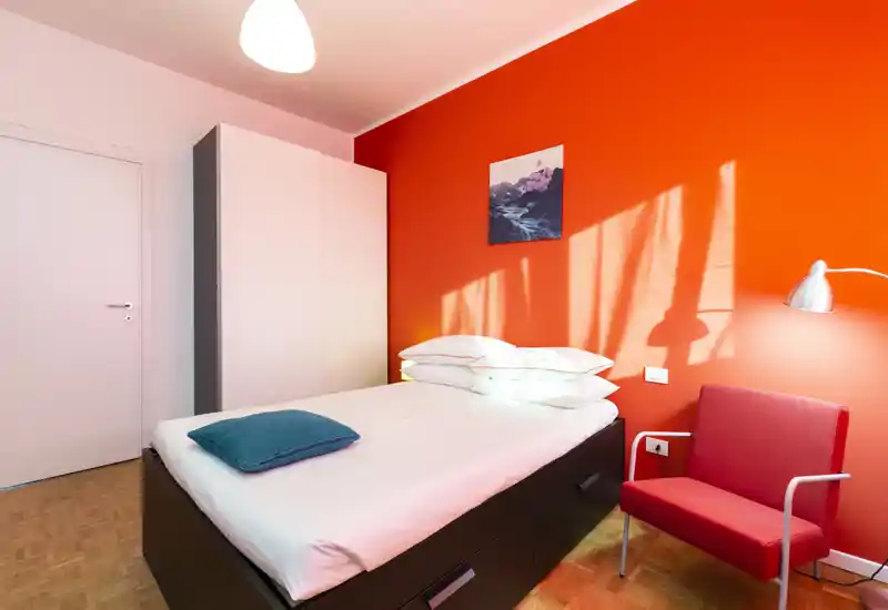 Camera singola moderna e luminosa con letto ben rifatto, parete arancione vivace e una comoda poltrona — ideale per soggiorni brevi.