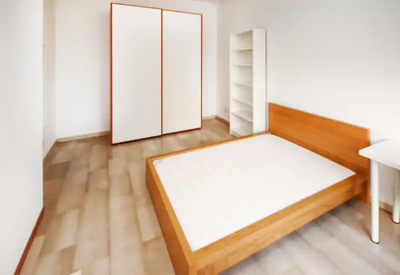 Dormitorio luminoso y minimalista con cama doble de madera, armario blanco y estantería; líneas limpias y suelo claro que crean un espacio sereno.