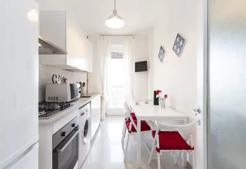 Cucina moderna a corridoio, luminosa, con elettrodomestici integrati, piccolo tavolo da pranzo e sedie con cuscini rossi vicino alla finestra soleggiata — perfetta per appartamenti compatti.