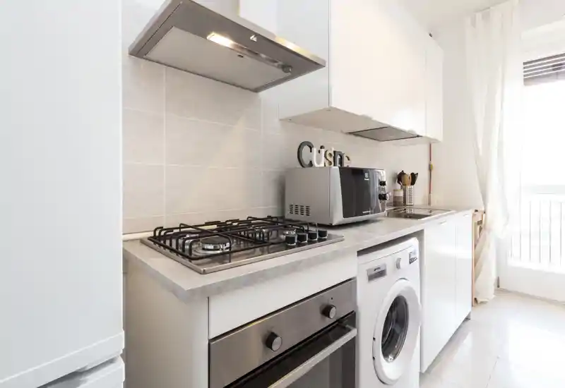 Cucina compatta, moderna e luminosa con piano cottura a gas, forno incassato, microonde e lavatrice sotto il piano; piastrelle chiare e finestra che donano un aspetto arioso.
