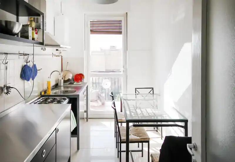 Cucina moderna e luminosa a corridoio con piani in acciaio inox, piano cottura a quattro fuochi, tavolo da pranzo in vetro e luce naturale dalla porta sul balcone.