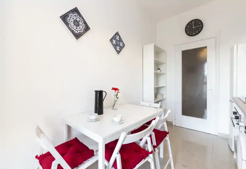 Angolo pranzo luminoso e minimal con tavolo bianco, quattro sedie con cuscini rossi, scaffale semplice e piastrelle decorative a parete — perfetto per colazioni accoglienti.