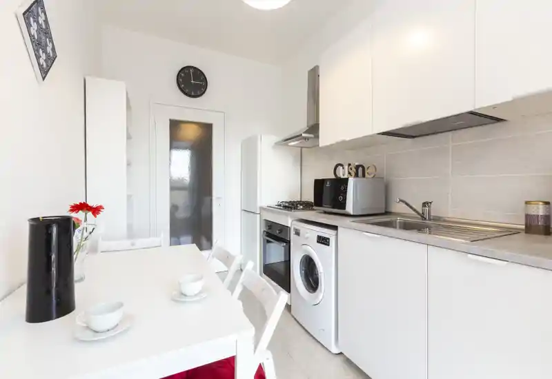 Cucina compatta e luminosa con mobili bianchi, forno integrato, lavatrice e piccolo tavolo da pranzo — stile moderno e pulito, perfetto per un appartamento in città.