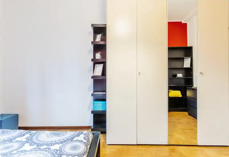 Camera da letto luminosa e ordinata con letto matrimoniale parzialmente inquadrato, un armadio alto con anta specchiata e mensole aperte con oggetti decorativi—buona luce naturale e stile curato.