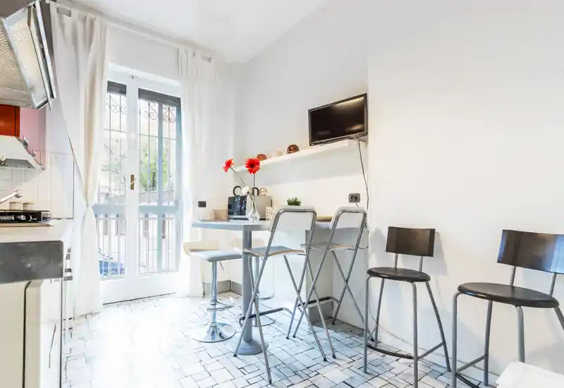 Angolo cottura moderno e luminoso con un piccolo bancone, sgabelli alti e una porta vetrata che lascia entrare molta luce naturale. Perfetto per appartamenti compatti in città.