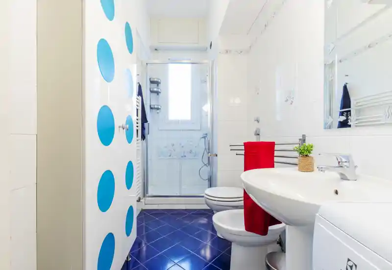 Bagno luminoso e moderno con piastrelle bianche, box doccia in vetro, bidet e WC, e un lavabo con un asciugamano rosso che crea contrasto.