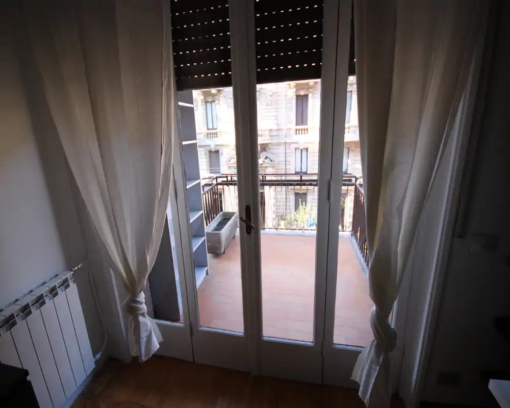 Piccolo balcone piastrellato accessibile tramite porte-finestre in vetro, incorniciato da tende leggere e con vista su una facciata cittadina classica — perfetto per un caffè mattutino.