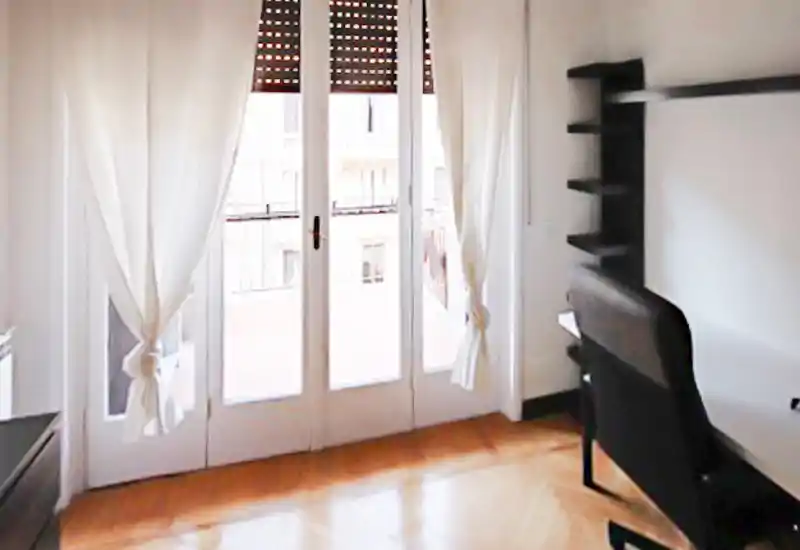 Stanza luminosa con grandi porte-finestre e tende leggere, pavimento in parquet e scrivania con sedia di lato — ideale come camera da letto o studio.
