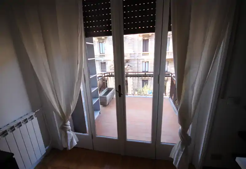 Piccolo balcone piastrellato accessibile tramite porte-finestre in vetro, incorniciato da tende leggere e con vista su una facciata cittadina classica — perfetto per un caffè mattutino.