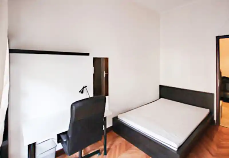 Camera da letto luminosa e minimal con letto matrimoniale, semplice testiera e una scrivania con sedia — pavimento in parquet e pareti neutre per un ambiente rilassante.