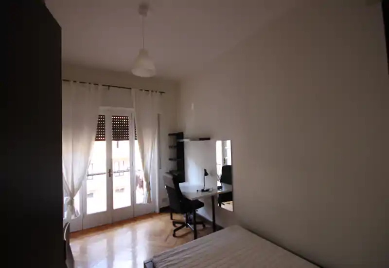 Camera da letto luminosa e arredata semplicemente con letto singolo, scrivania e porta balcone che illumina l'ambiente. Pavimento in parquet e pareti neutre rendono la stanza versatile.