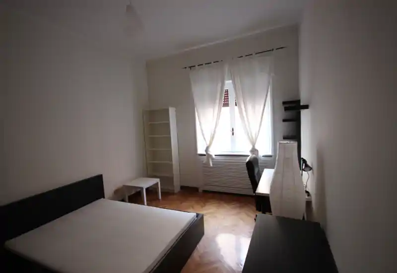 Camera da letto minima e luminosa con letto matrimoniale, pavimento in parquet, scrivania vicino alla finestra e mensole semplici. Ampia finestra con tende leggere che diffondono luce naturale.