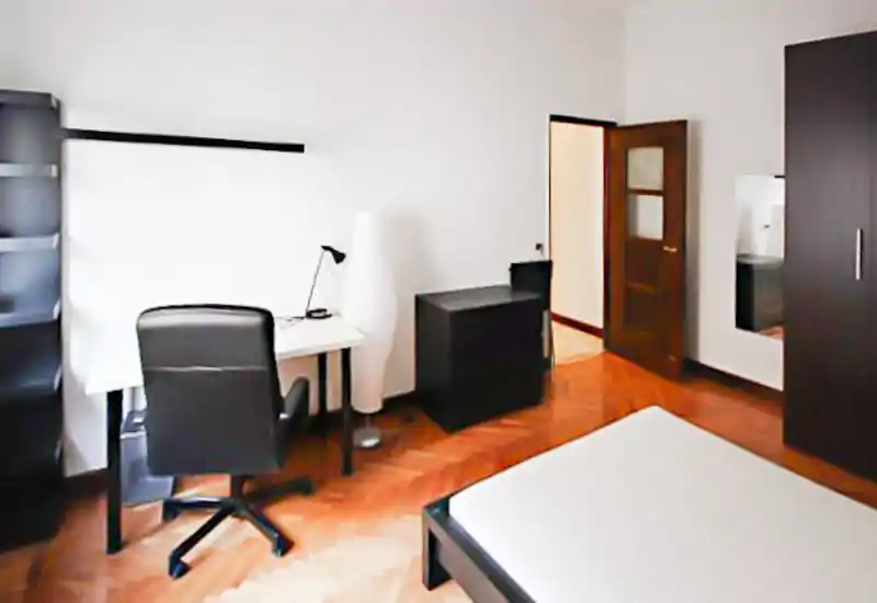 Camera da letto luminosa e arredata in modo semplice con letto matrimoniale visibile, scrivania e armadio; parquet in legno e mobili scuri minimal creano uno spazio pulito e funzionale ideale per affitti brevi.