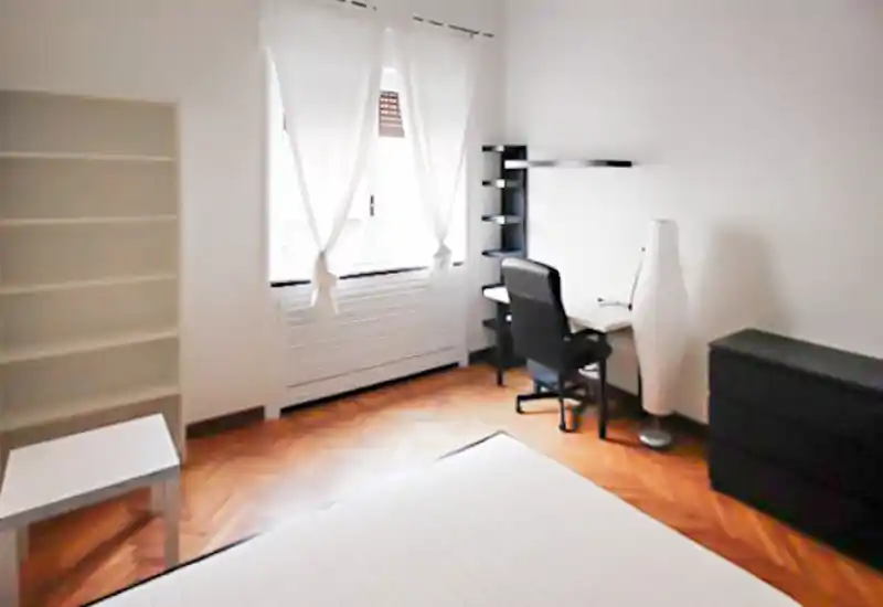 Camera da letto luminosa e minimale con materasso in primo piano, scrivania vicino alla finestra, scaffale e comò — linee pulite e molta luce naturale.