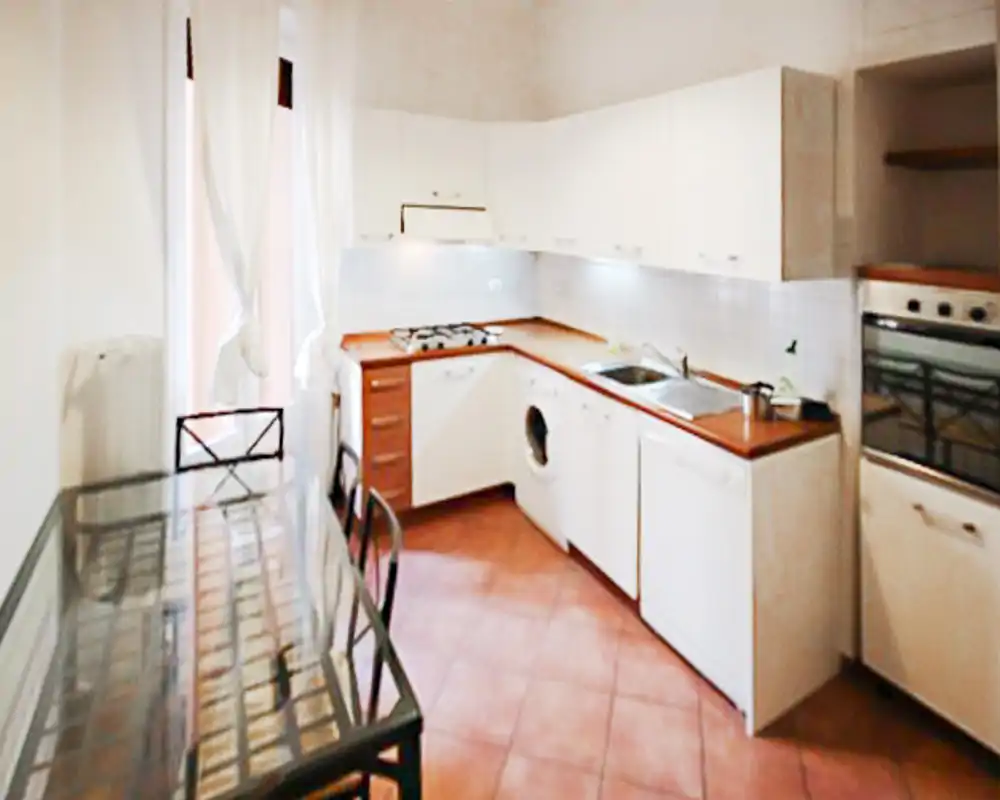 Cucina compatta e luminosa con mobili bianchi a L, top in legno, forno incassato e tavolo da pranzo in vetro; pavimento in cotto e luce naturale creano un ambiente accogliente.