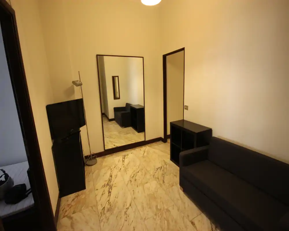 Soggiorno compatto con divano scuro, scaffalatura bassa e un grande specchio a parete che amplia visivamente lo spazio; illuminazione calda dall'alto e pavimento in marmo per un aspetto essenziale.