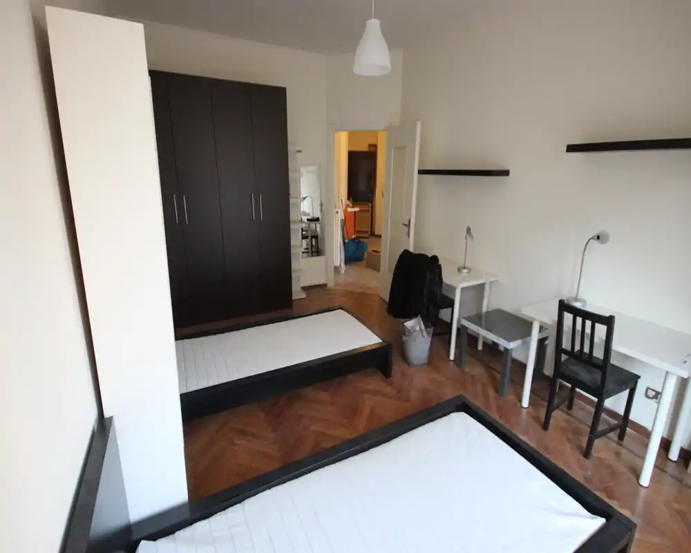 Spacious twin-bedroom with two single beds, a large dark wardrobe and two desks — bright room with parquet flooring and simple, modern furnishings.