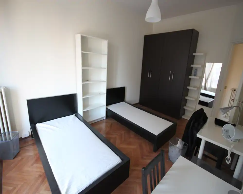 Bright twin-bedroom with two single beds, dark wooden frames and parquet flooring. The room includes a large wardrobe, shelving, desk and natural light from a side window — practical and ready for renters.
