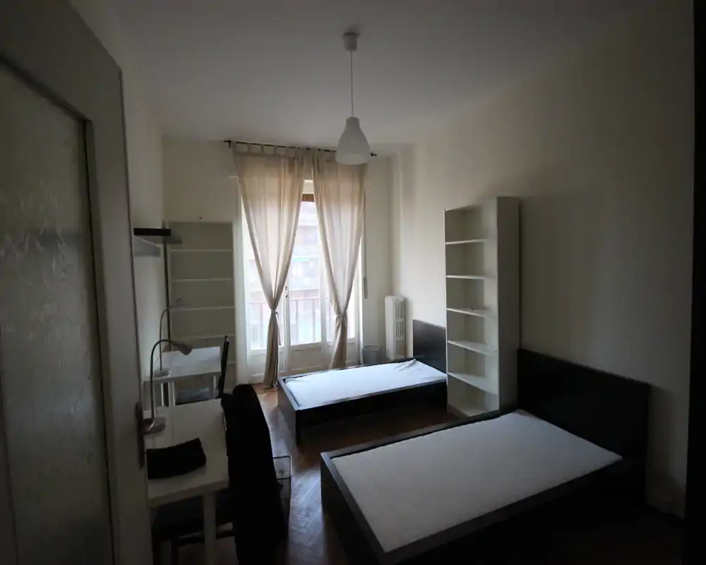 Bright twin-bedroom with two single beds, tall window with sheer curtains and simple shelving units — ideal for shared students or short-term stays.