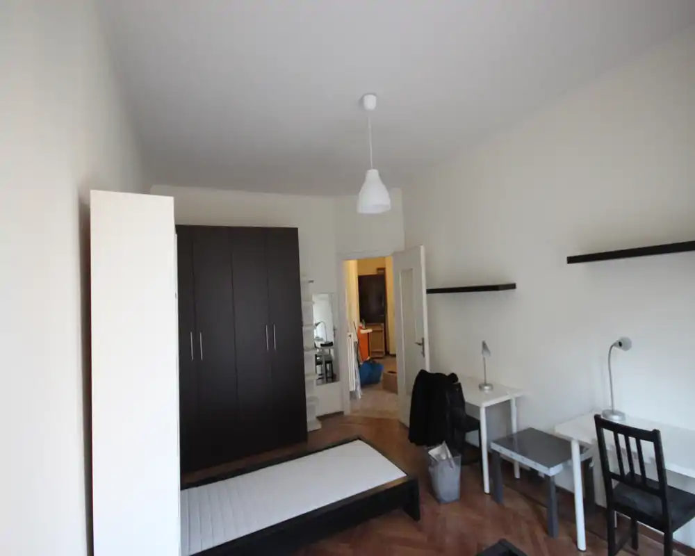 Simple, bright bedroom with a single bed frame and mattress, dark wardrobe, desk and shelving — clean, minimalist layout ideal for a student or single occupant.