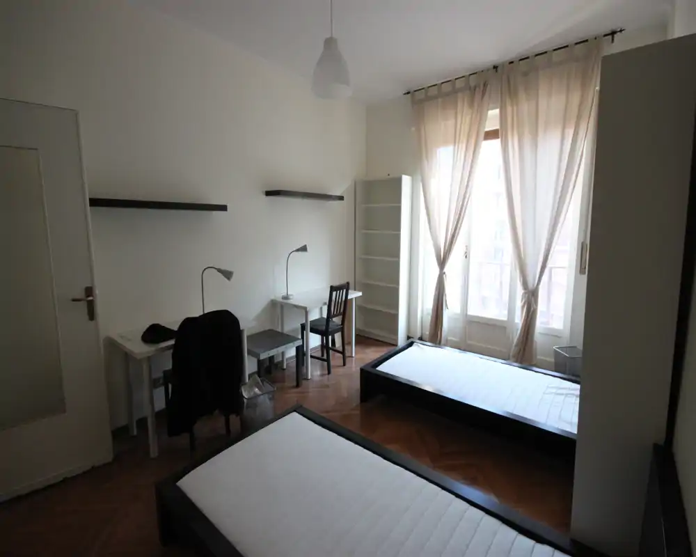 Bright twin-bedroom with two single beds on low black frames, parquet floor and large French windows with sheer curtains letting in soft natural light. Two small desks, shelving and minimal decor make it a practical shared student or guest room.