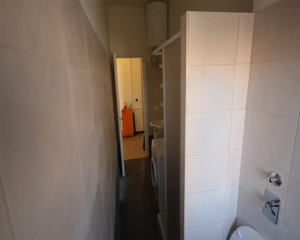 Narrow bathroom with tiled walls, a shower stall, sink and toilet visible; compact, functional layout with neutral tiles and natural light from the doorway.