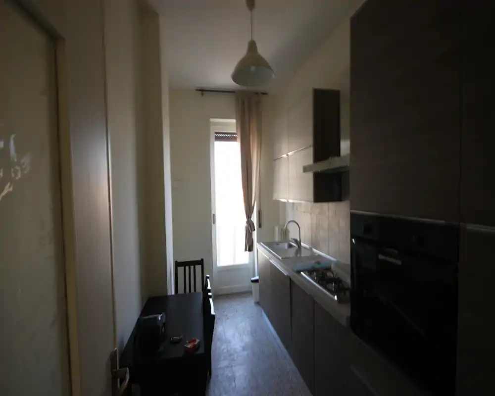Compact galley kitchen with modern wood-effect cabinets, integrated oven, sink and gas hob; a small table near the entrance and a glass door provide natural light.