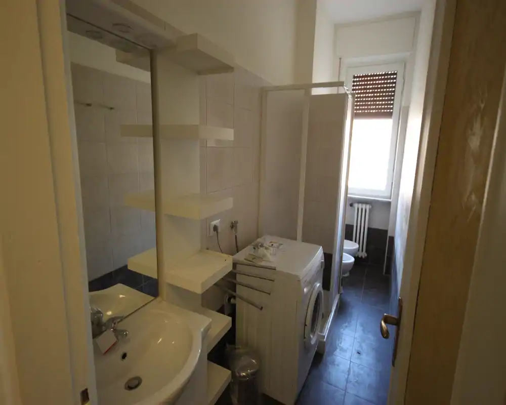 Small bathroom with sink, mirror cabinet, shower stall, washing machine and toilet visible near a window; compact, functional layout.