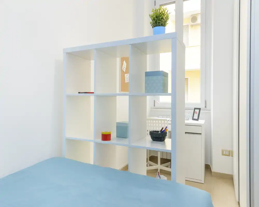 Dormitorio luminoso y minimalista con colcha azul y estantería de cubos que divide el espacio; luz natural desde la ventana y cajas decorativas que aportan sensación ordenada y fresca.