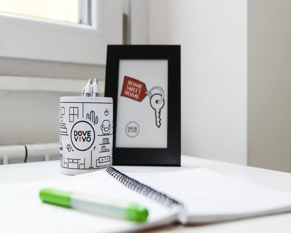 Rincón de sala luminoso y minimalista con un marco decorativo y un portalápices de marca sobre un escritorio blanco, fotografiado con poca profundidad de campo.