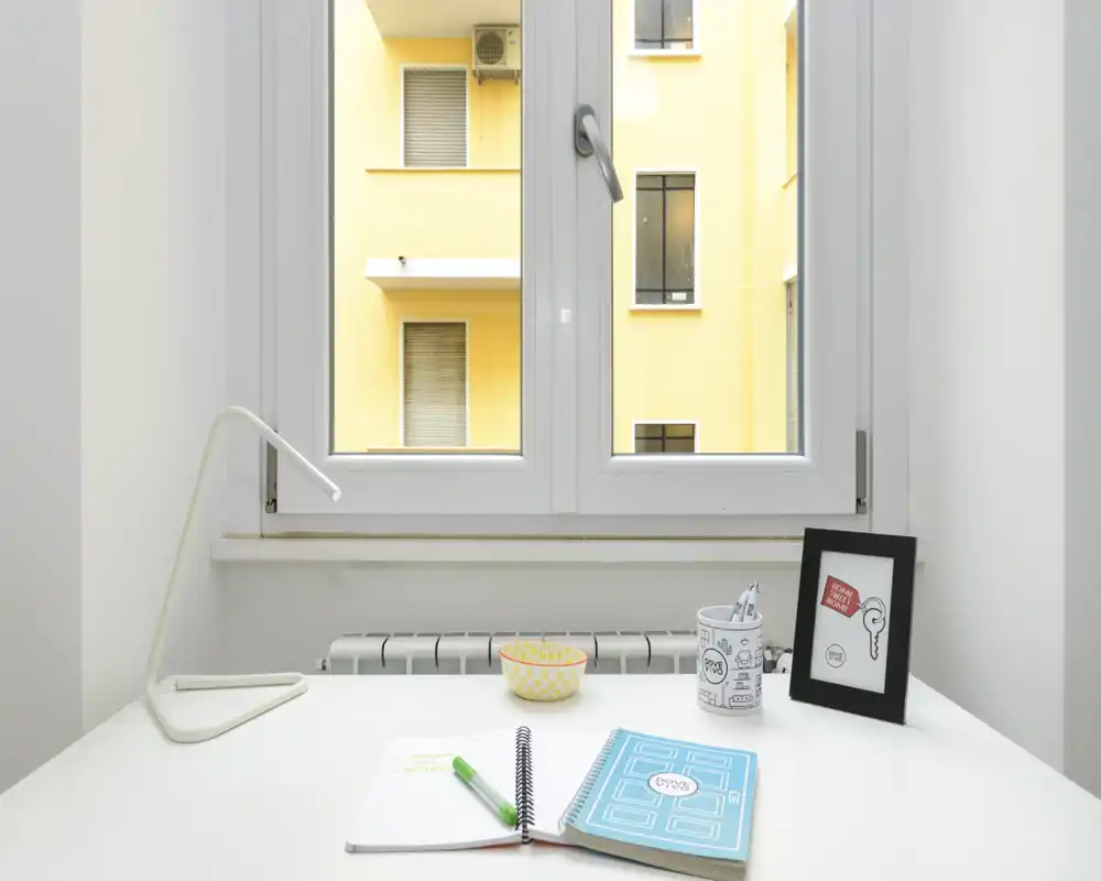 Área de escritorio luminosa y minimalista junto a una gran ventana, con lámpara, libreta, bolígrafo, bol y marco. La luz natural inunda el espacio, ideal para trabajar o estudiar.