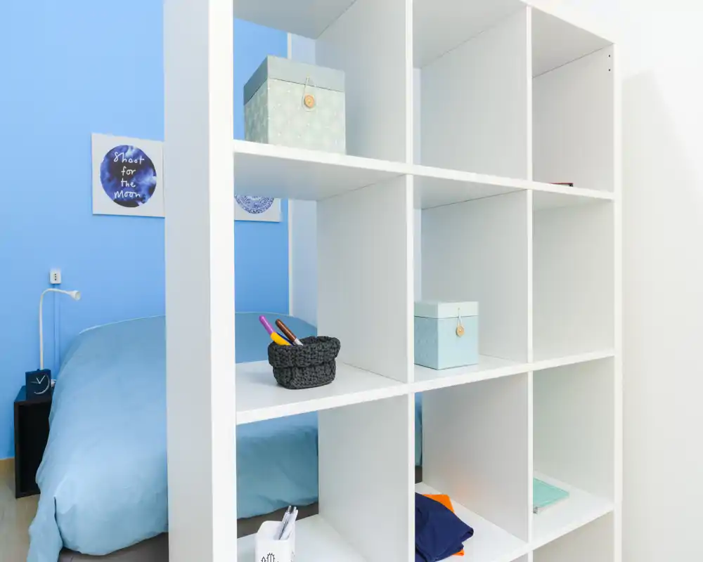 Dormitorio luminoso y moderno visto a través de una estantería de cubos blanca; paredes azul claro y ropa de cama a juego crean una sensación fresca y aireada con almacenamiento decorativo ordenado.