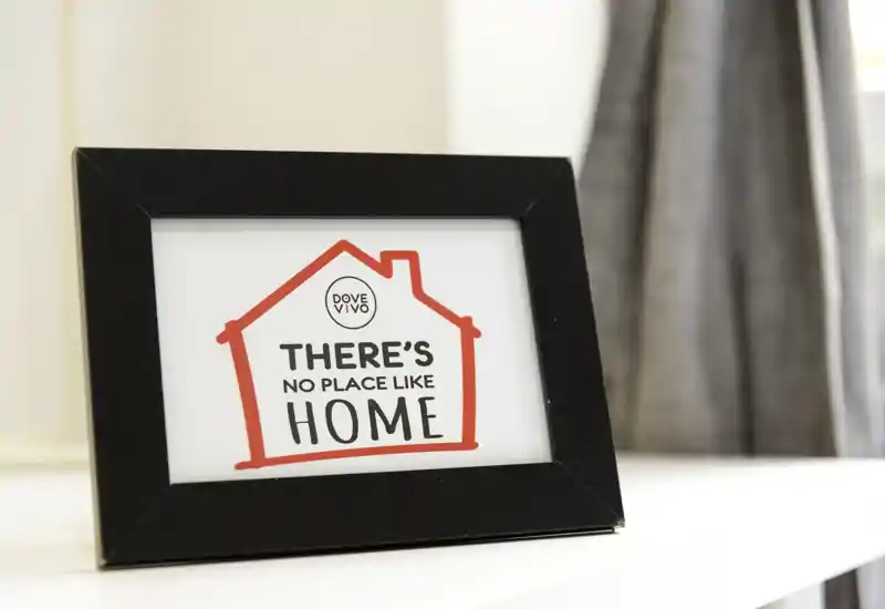 Close-up of a decorative framed sign that reads "There's no place like home" placed on a flat surface; a curtain is visible in the soft-focus background. The photo highlights decor detail rather than a room layout.