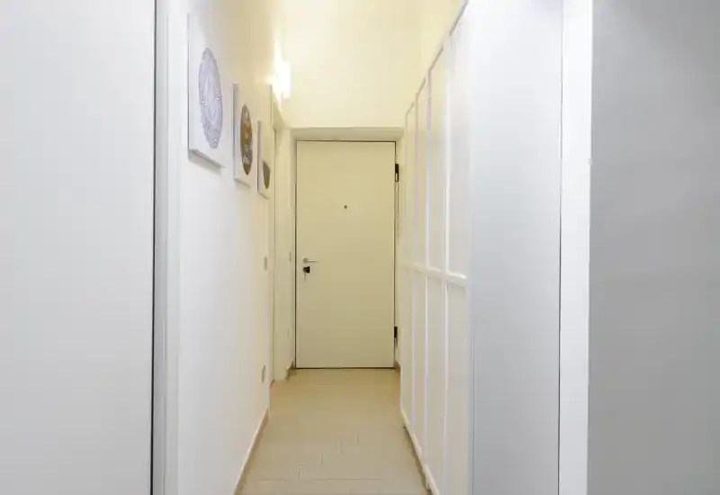 Bright, minimal entrance corridor with a white front door, light wooden floor and simple wall decor — clean and welcoming access to the apartment.