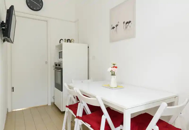 Bright, compact dining area with a white table and four chairs dressed with red cushions, a small vase of flowers and a clean minimalist décor — ideal for casual meals.