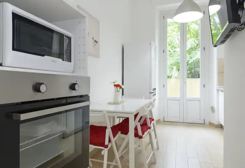 Bright, modern kitchen with integrated oven, microwave and a small dining table with red-cushioned chairs; large window/door brings abundant natural light.