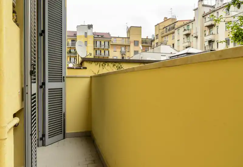 Small private balcony with yellow walls overlooking neighboring buildings; tiled floor and shuttered door provide easy access and privacy.