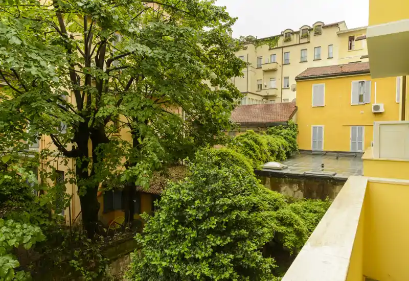 View from a balcony overlooking a lush courtyard with mature trees and yellow-painted surrounding buildings; bright, leafy urban outdoor space.