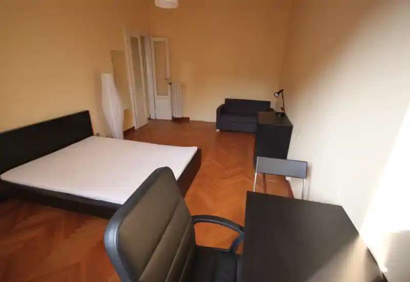 Ampia camera da letto con letto matrimoniale, scrivania e sedia semplici, un piccolo divano e pavimento in parquet caldo. Luce naturale abbondante e arredi moderni e minimali, ideale per annunci in affitto.