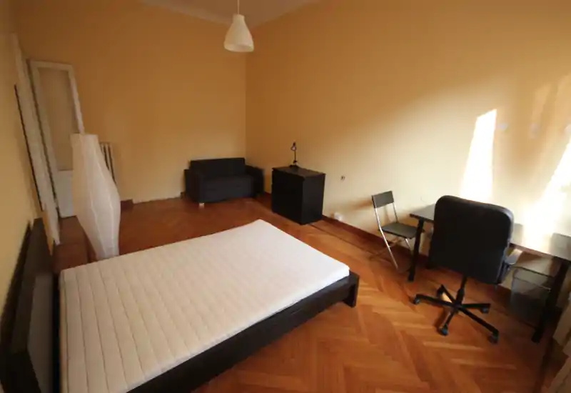 Camera da letto luminosa e poco arredata con letto matrimoniale, scrivania e sedia vicino alla finestra, piccolo divano e pavimento in parquet. La luce naturale illumina la stanza donando un'atmosfera calda.