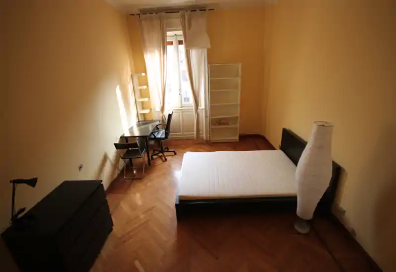 Ampia camera da letto con letto matrimoniale su struttura bassa, pavimento in parquet e angolo finestrato luminoso con scrivania e scaffali — arredo semplice, caldo e ben illuminato.