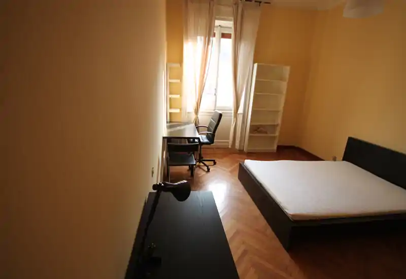 Camera da letto luminosa e minimale con letto matrimoniale su struttura bassa, pavimento in parquet e una scrivania vicino alla finestra con sedia. Tendine leggere diffondono la luce naturale e una libreria bianca offre spazio contenitivo.