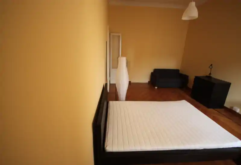 Camera da letto luminosa e poco arredata con un grande letto basso, pavimento in parquet, una lampada da terra e un piccolo angolo salotto.