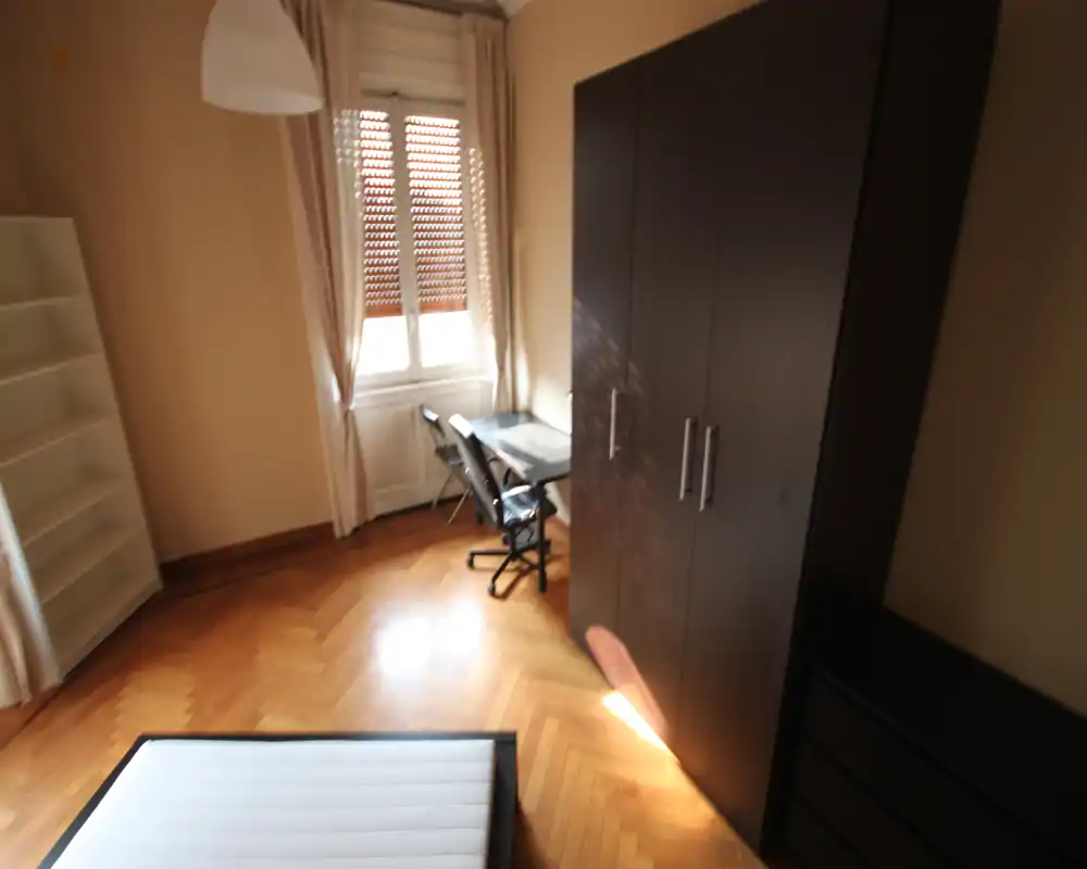 Camera da letto con materasso in primo piano, pavimento in parquet, grande armadio scuro e scrivania vicino alla finestra che lascia entrare luce naturale.