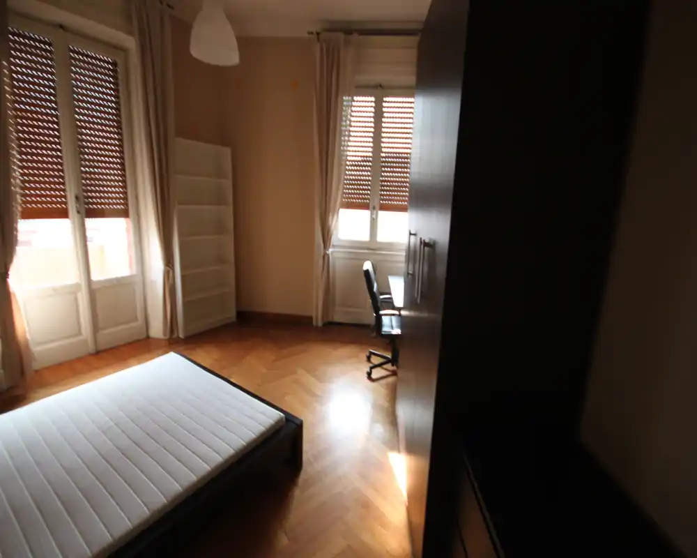 Camera da letto luminosa con struttura letto e materasso visibili, pavimento in parquet, grandi finestre con tende e un armadio; ambiente semplice e arioso ideale per annunci.