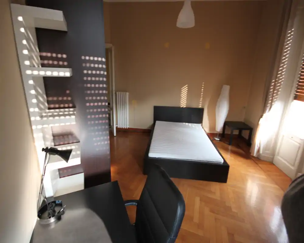 Camera da letto luminosa e poco arredata con struttura del letto matrimoniale, scrivania e pavimento in parquet. Ampia finestra che dona luce naturale calda, arredi moderni e una lampada da terra.