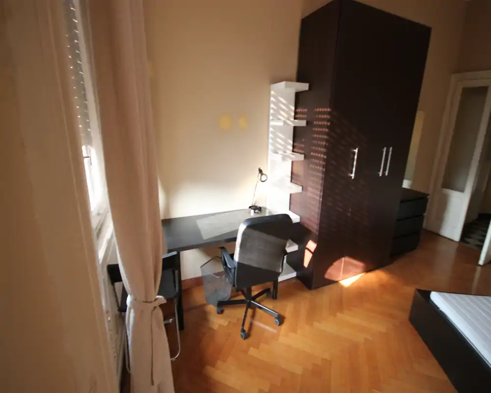Angolo di camera luminoso e arredato con letto parzialmente visibile, scrivania e sedia vicino alla finestra e un armadio alto scuro con ampio spazio di contenimento.