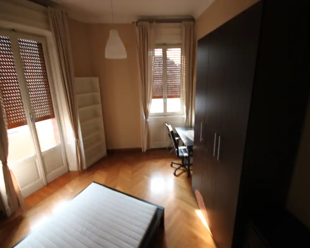 Camera da letto luminosa con materasso su struttura bassa, grande armadio, scrivania e luce naturale da due finestre con tende.