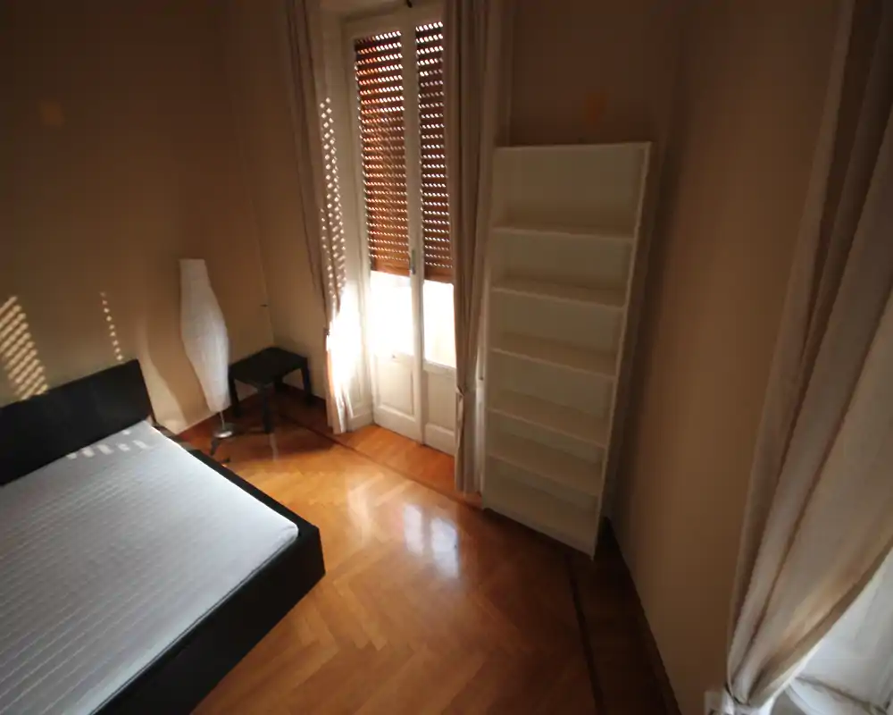 Camera da letto luminosa con letto basso moderno, pavimento in parquet, finestra alta con persiane e tende leggere, scaffale e lampada indipendenti — spazio semplice e arioso con molta luce naturale.